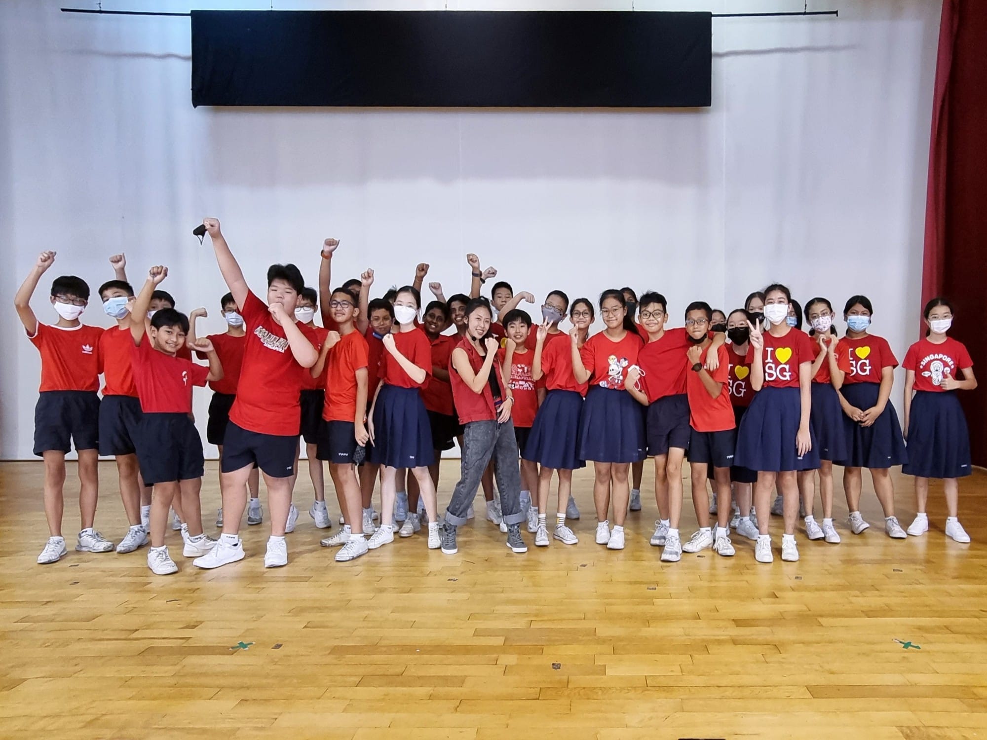 A large group of students in red shirts posing with raised fists on a wooden hall floor.
